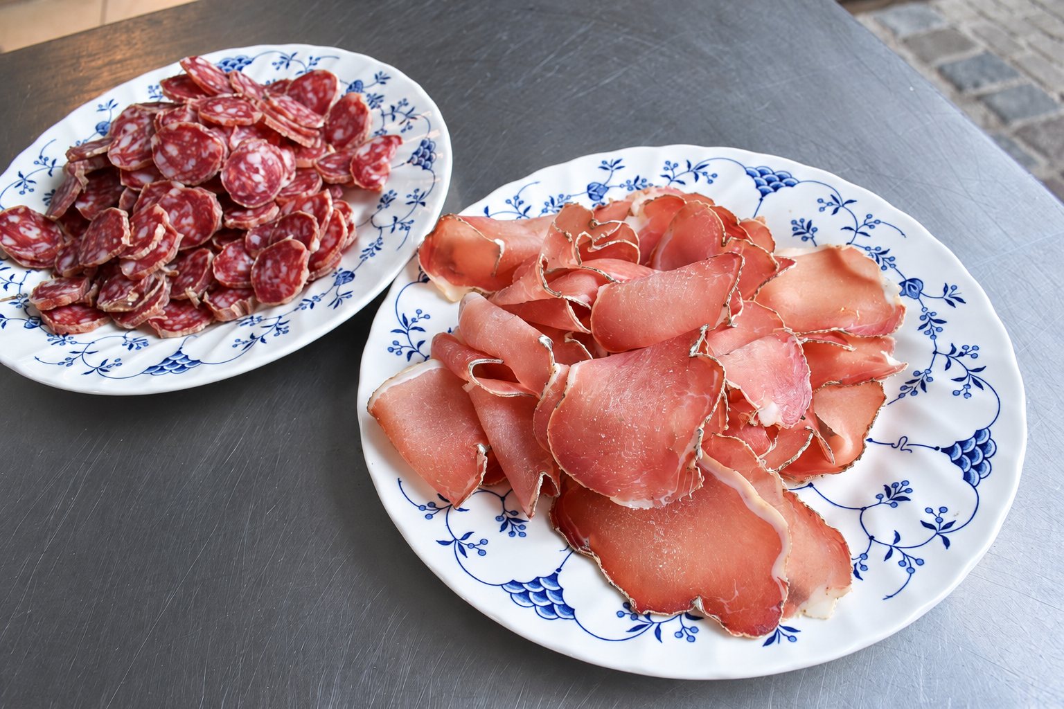 Deux assiettes en porcelaine blanche à motifs bleus posées sur une table en métal, l’une garnie de rondelles de saucisson sec et l’autre de fines tranches de jambon cru.