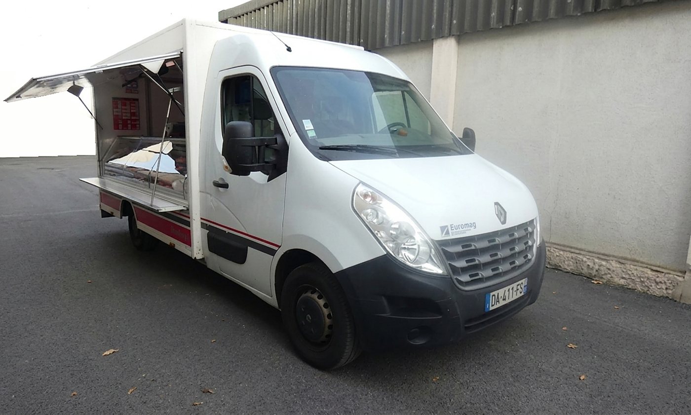 Un camion magasin blanc de marque Renault, volet latéral relevé et vitrine réfrigérée visible, stationné devant un mur gris.