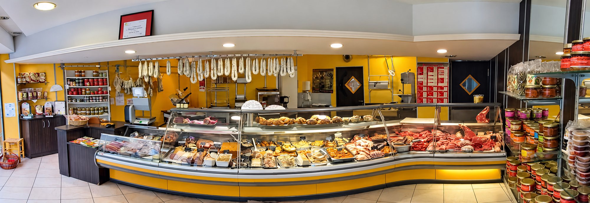 Vue panoramique de l’intérieur d’une boucherie-charcuterie aux murs jaunes, avec une vitrine réfrigérée garnie de viandes et de plats préparés, des saucisses suspendues et des bocaux sur les étagères.