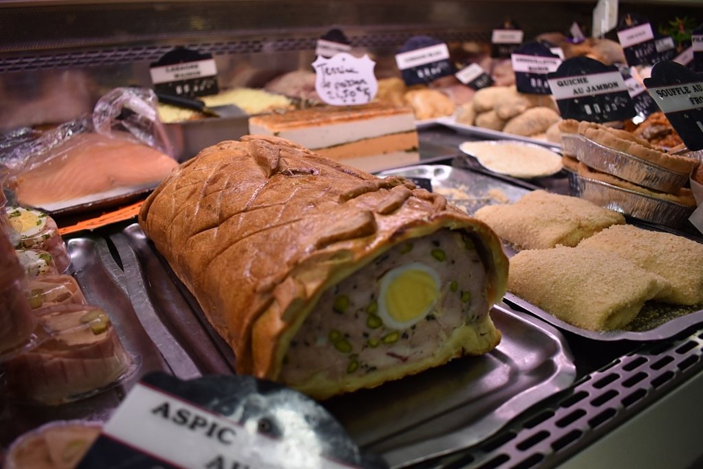 Une tranche de pâté en croûte garni d’un œuf dur et de pistaches est présentée dans la vitrine d’un traiteur, entourée de quiches et d’autres préparations.