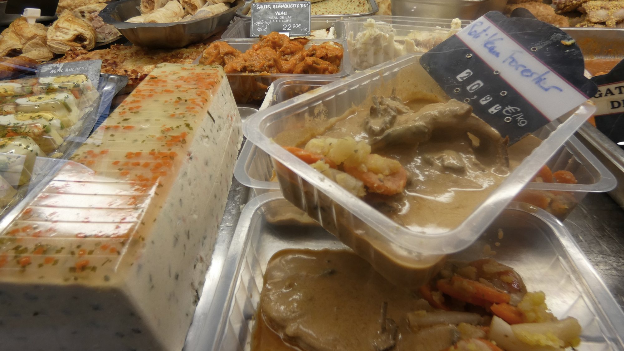 Vitrine de traiteur présentant des barquettes de viande en sauce avec légumes, une terrine en tranches et des étiquettes de prix.