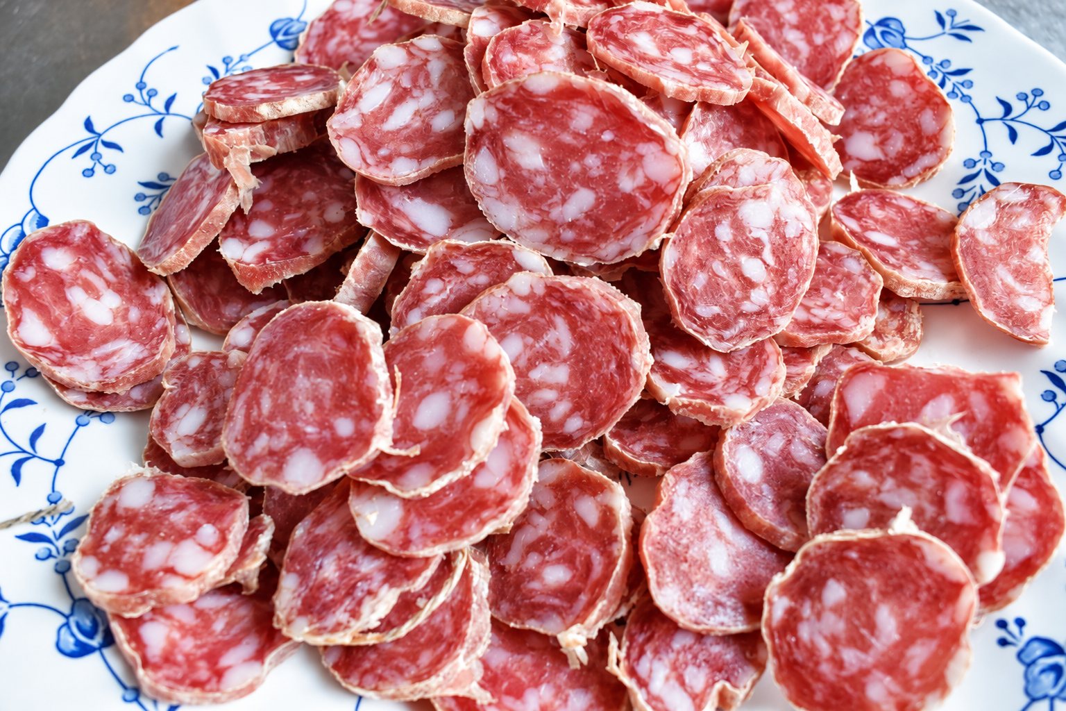 Un tas de fines rondelles de saucisson sec posées sur une assiette blanche décorée de fleurs et de feuilles bleues.