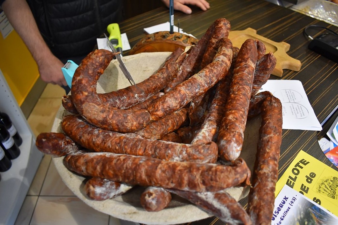 Un tas de saucisses sèches rougeâtres posées sur un grand plateau rond, sur un comptoir en bois avec des prospectus et un vendeur en arrière-plan.