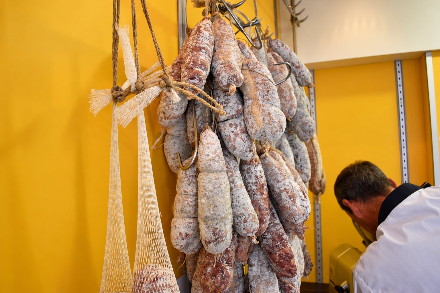 Des saucissons secs à la croûte blanche sont accrochés à des crochets près de filets de boucher, tandis qu’un homme en veste blanche se penche vers une trancheuse au second plan.