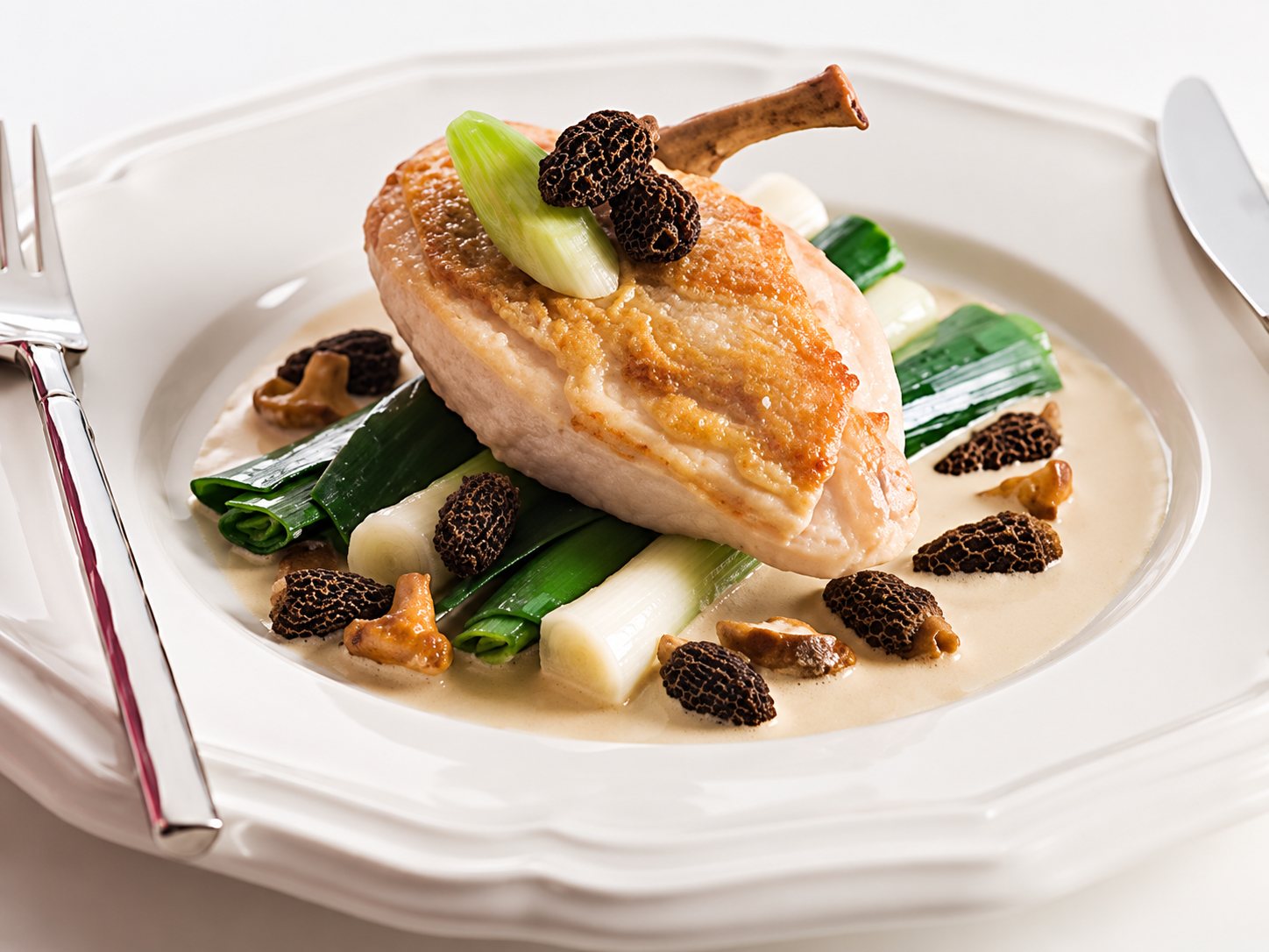 Assiette blanche garnie d’une volaille rôtie dorée, de morilles et de poireaux braisés dans une sauce crémeuse.