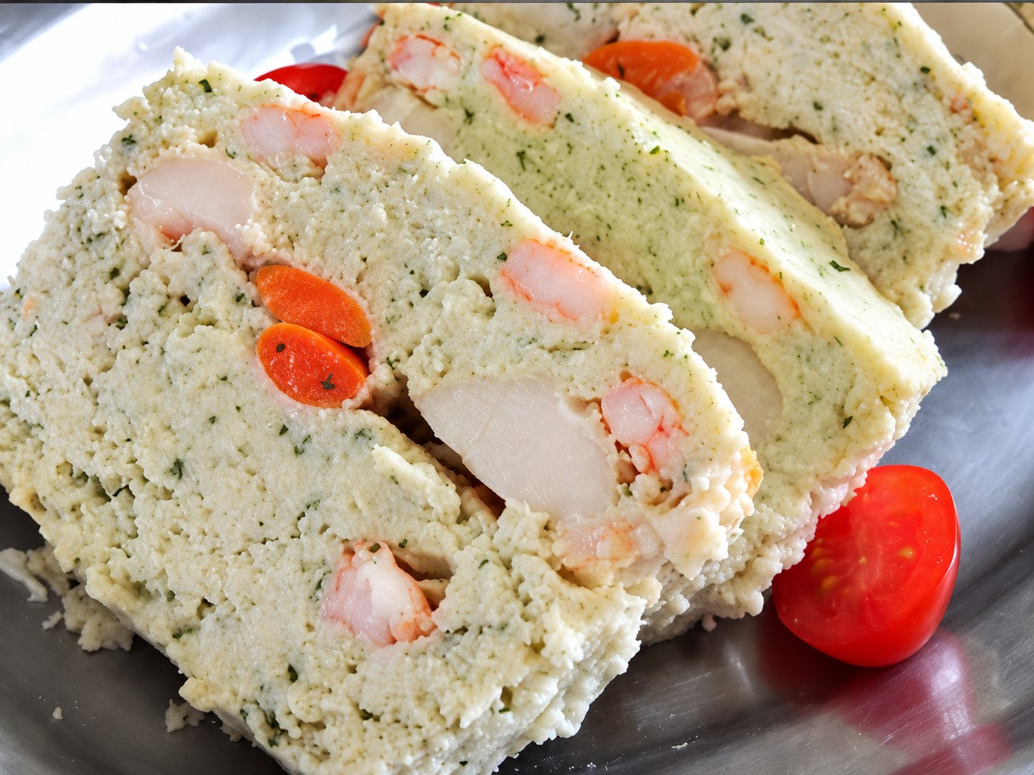 Gros plan sur des tranches de terrine blanche aux herbes garnie de morceaux de crevettes et de carottes, accompagnées de tomates cerises sur un plat métallique.