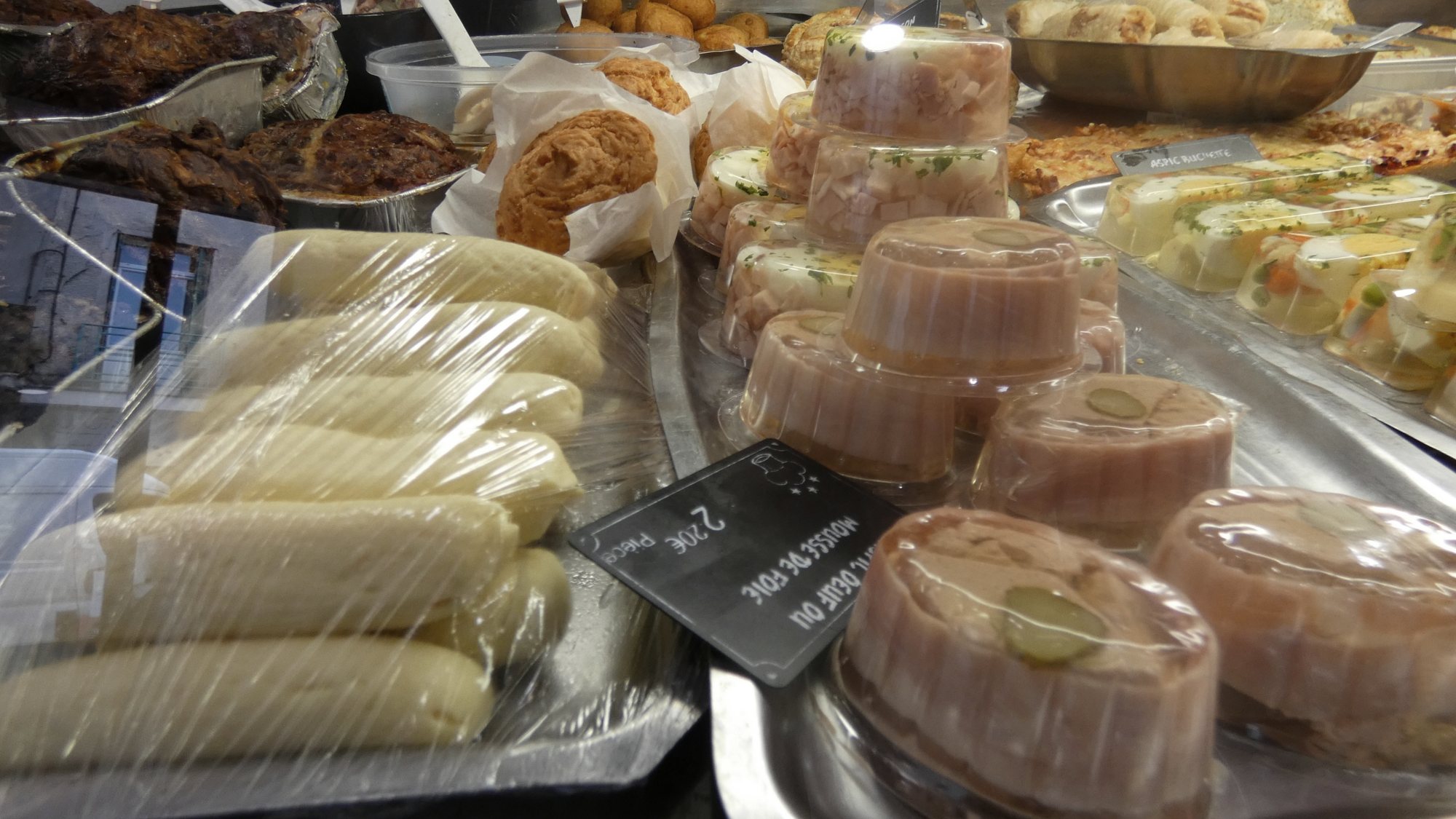 Vitrine de traiteur présentant des boudins blancs sous film, des terrines de jambon en gelée, des brioches et une étiquette de mousse de foie.
