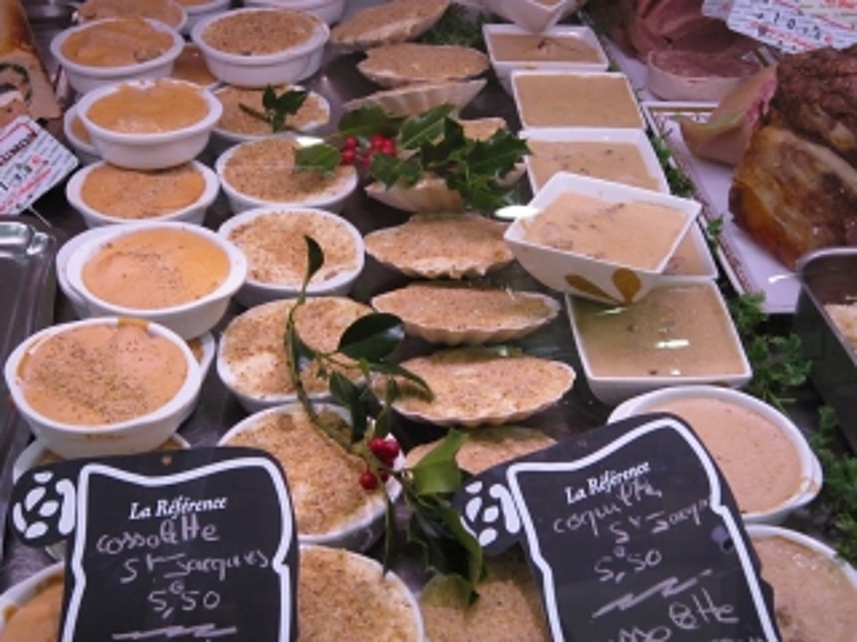 Une vitrine de traiteur présente des coquilles Saint-Jacques gratinées et des ramequins de préparations, avec des ardoises de prix décorées de houx.
